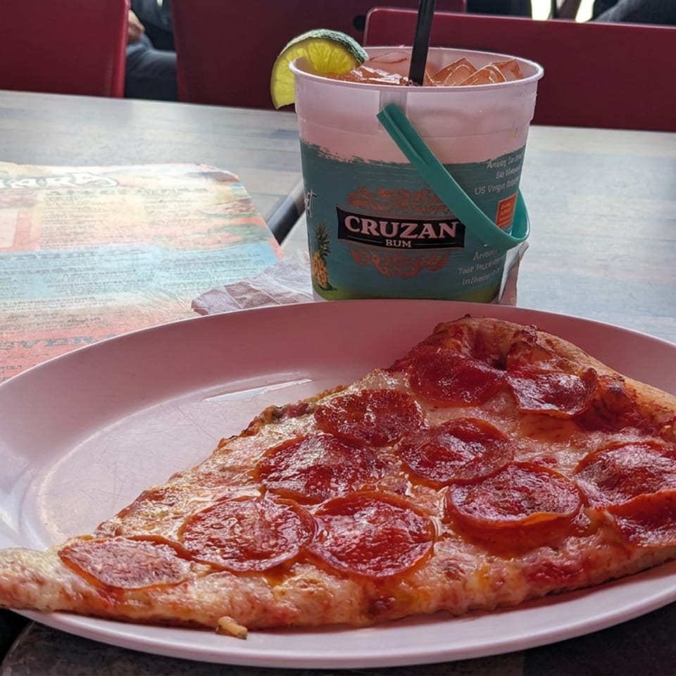 Large pepperoni pizza slice on a white plate beside an iced Cruzan Rum drink, menu, and red restaurant chairs.