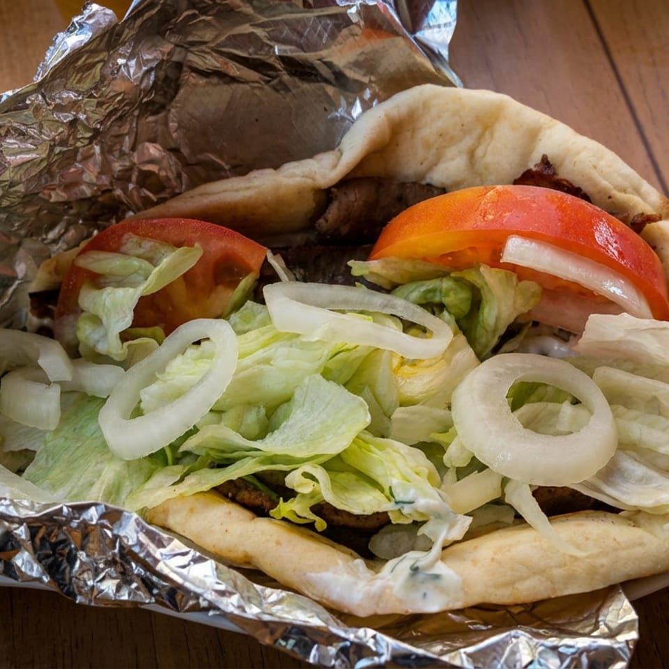 Open pita sandwich filled with grilled meat, lettuce, tomatoes, onions, and creamy sauce on foil-lined plate.
