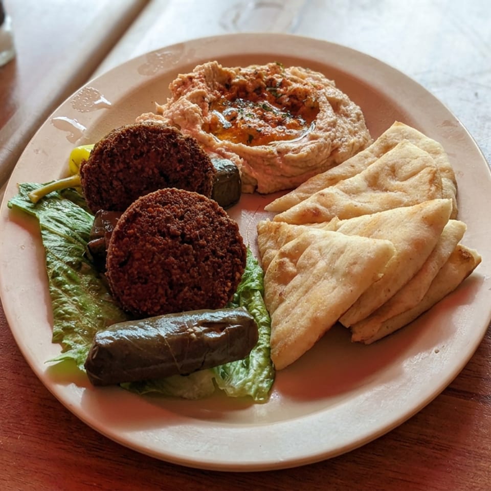 Mediterranean platter with falafel, hummus, grilled sausage, pita bread, and greens on a wooden restaurant table.